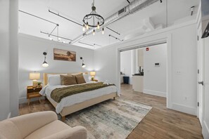 Ein modernes Schlafzimmer mit einem großen Bett, Nachttischlampen, einer Chandelierleuchtre und Blick in die angrenzende Küche.
