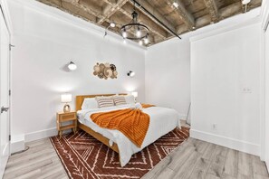 A bedroom with a bed, bedside lamps, a rug, and a ceiling with exposed beams. - NYC 20 Min Designer Loft | Gym, Desk & Parking (Kearny)