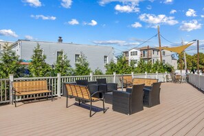 A wooden deck with outdoor seating, a white fence, and buildings in the background. - NYC 20 Min Designer Loft | Gym, Desk & Parking (Kearny)