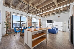 A modern kitchen with a central island, dining area with chairs, a sofa, and a TV mounted on the wall. - 20 Min NYC Brick Loft w/ Gym, Desk & Parking (Kearny)