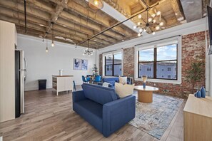 A spacious living area with a blue sofa, dining table, and chairs, a kitchen area with a refrigerator, and large windows offering views of buildings. - 20 Min NYC Brick Loft w/ Gym, Desk & Parking (Kearny)