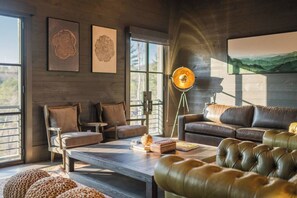 Ein modernes Wohnzimmer mit Ledersofa, Couchtisch und Stehlampe. An den Wänden hängen gerahmte Kunstwerke und große Fenster bieten einen Blick ins Freie.