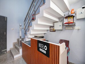 Ein Hotel-Rezeptionstresen mit einem Schild, das eine betriebliche Abwicklung anzeigt, eine Treppe mit Metallgeländer und ein an der Wand befestigtes Regal mit einem gerahmten Bild und einer kleinen Statue.