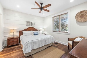 Ein Schlafzimmer mit einem Bett, einem Nachttisch, einer Lampe, einem Deckenventilator, einem Fenster mit Jalousien und einem Korbstuhl.