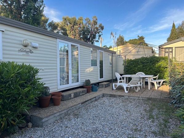 A mobile home with a patio area, including a table and chairs, surrounded by potted plants and a gravel driveway.