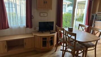 Ein Zimmer mit einem Holzschrank, einem Fernseher, einem Esstisch mit Stühlen und einem Fenster mit Vorhängen.