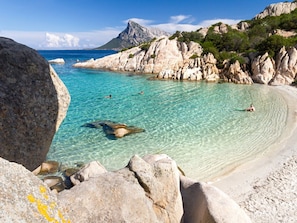 Ein Küstenstrand mit klarem türkisblauem Wasser, Felsformationen und einem Berg im Hintergrund.