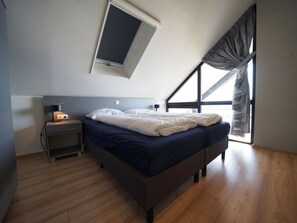 Ein Schlafzimmer mit schrägem Dach, einem Bett, einem Nachttisch, einer Lampe und einem Fenster mit Vorhängen.