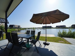 Eine Terrasse im Freien mit Tisch und Stühlen, einem Sonnenschirm und Blick auf ein Gewässer mit Gebäuden im Hintergrund.