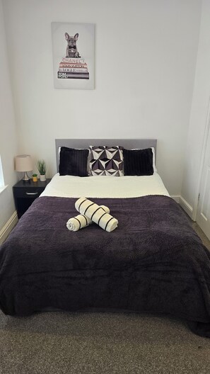 A neatly made bed with a dark purple blanket and two pillows, a small decorative item on the nightstand, and a framed picture on the wall. - 4 Bedroom House all En-suite rooms. (Stoke-on-Trent)