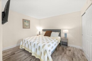 A bedroom with a bed, bedside lamps, a nightstand, a wall-mounted TV, and a door. - Xbeach Villa 2516-oop Honor Bookings 1 Bedroom Condo (Captiva)