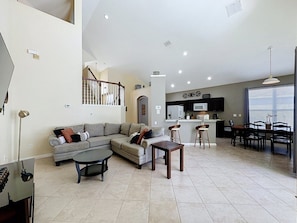 A spacious living area with a sofa, coffee table, and dining table. A kitchen with a microwave and bar stools is visible in the background. - Luxury 5bd 3.5ba Pool Home In Windsor Palms Resort 5 Bedroom Villa (Kissimmee)