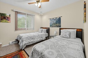 Habitación de hotel con dos camas, ventilador de techo, ventana con persianas y cuadros en las paredes.
