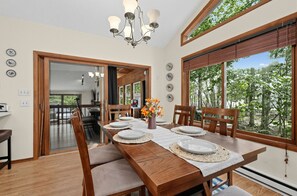 A dining area with a wooden table set for four, a chandelier, and large windows with a view of trees. - Ski-in Ski-out Camel Back 10min Big Boulder 15min 4 Bedroom Home (Tobyhanna)