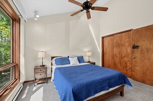 A bedroom with a bed, two bedside tables, a ceiling fan, a wardrobe, and a window with a view of greenery.