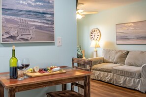 Ein Wohnzimmer mit einem Holztisch, der für eine Mahlzeit gedeckt ist, einer Couch, einer Uhr und einem Gemälde mit Strandmotiv.