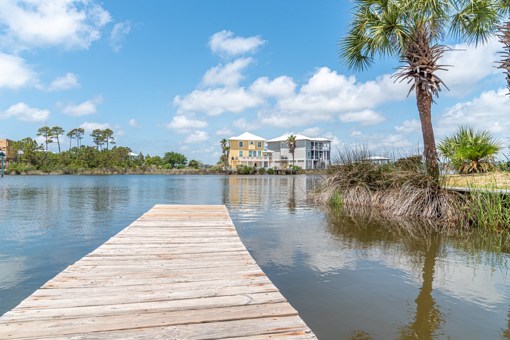 A wooden dock leading to a waterfront property with multiple buildings, a palm tree, and a clear sky.