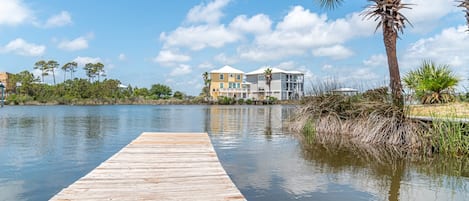 A wooden dock leading to a waterfront property with multiple buildings, a palm tree, and a clear sky.