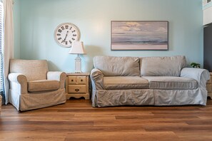 A living room with a beige sofa, a matching armchair, a side table with a lamp, a wall clock, and a landscape painting. - The Cove 104b 1 Bedroom Condo (Gulf Shores)