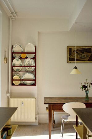 Een kamer met een houten tafel, een witte stoel, een radiator en een wandplank met borden.