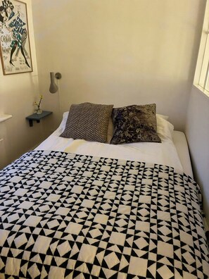 Een bed met een beddekleed met een geometrisch patroon, twee kussens met verschillende designs, een wandlamp en een inlijsting foto aan de muur.