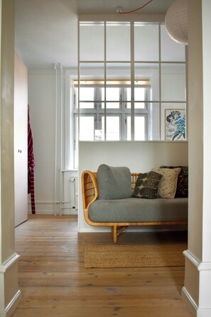 A room with a wooden floor, a grey sofa with cushions, a wicker chair, and a window with blinds. - Newly renovated flat with balcony in Nørrebro (København)