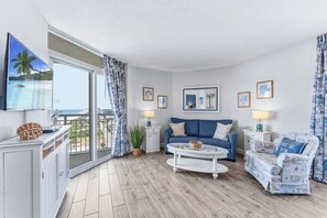 Un salon avec un canapé, une table basse et un fauteuil, un balcon avec vue et un téléviseur fixé au mur.