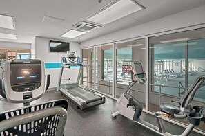 Une salle de sport équipée de tapis de course, de vélos d’exercice et offrant une vue sur la piscine.
