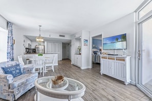 A modern living room with a round glass table, a sofa with ocean-themed cushions, and a television on a cabinet. - Bay Watch Condo with floor to ceiling windows | Bay Watch Resort - 1641 (North Myrtle Beach)