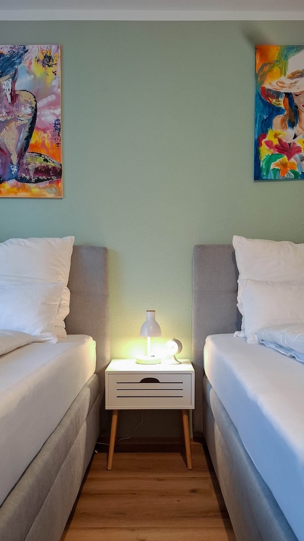 A bedroom with two beds, a bedside table with a lamp, and two paintings on the wall.