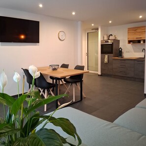 A modern dining area with a wooden table and black chairs, a clock on the wall, and a kitchen in the background.