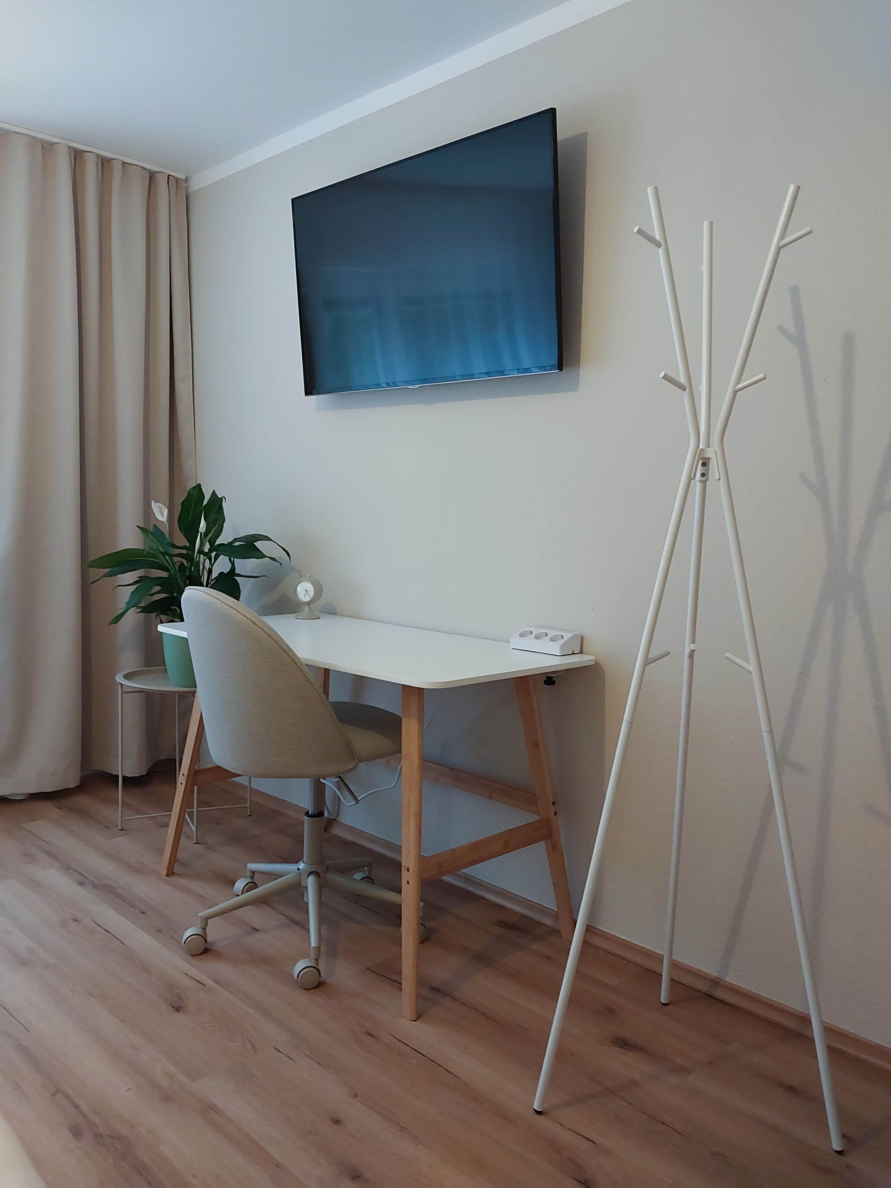 Ein Zimmer mit Schreibtisch, Stuhl, Topfpflanze und Kleiderständer.