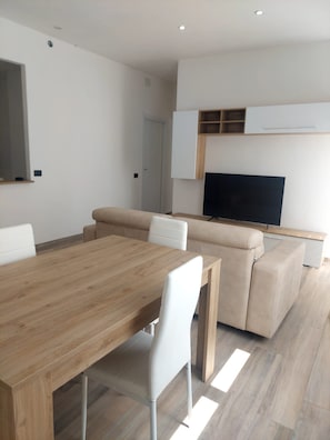A modern living room with a wooden dining table, white chairs, a beige sofa, and a flat-screen TV mounted on the wall. - B&B del CorsoB&B del Corso (Perdasdefogu)