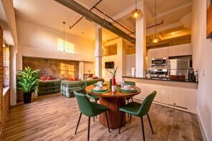 Eine moderne Küche mit Edelstahlgeräten, ein Essbereich mit einem runden Holztisch und grünen Stühlen sowie eine gemütliche Sitzecke mit Backsteinwand und grünen Sofas.