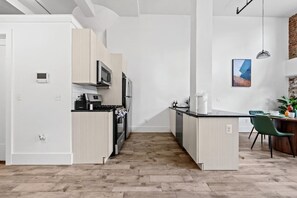 A modern kitchen with a central island, stainless steel appliances, and a dining area with a green chair. - The Garden Loft Near NYC + Parking + Patio (Kearny)