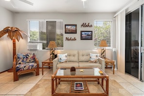 A living room with a sofa, two armchairs, a coffee table, and a TV. There are two air conditioning units, a window with blinds, and a wall with two framed pictures.
