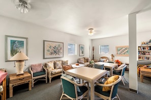 A spacious living area with a dining table, comfortable seating, and a bookshelf filled with books. - Makolea#21: Steps from the Beach, Beautifully Appointed 2 bedroom condo (KAILUA KONA)