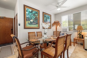 A dining area with a glass table, wicker chairs, and two framed tropical paintings.