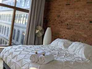 A hotel room with a brick wall, a large window, a bed with a geometric-patterned bedspread, two rolled towels, a standing fan, and a white pillow. - Stunning condo with a view, parking, and fireplace (Québec)