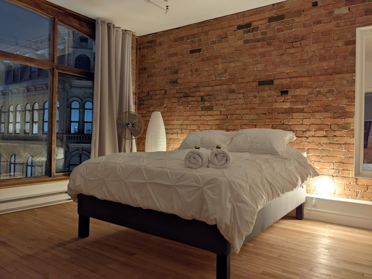 Una camera d'albergo con una parete in mattoni, un'ampia finestra, un letto con biancheria bianca e una lampada da comodino.