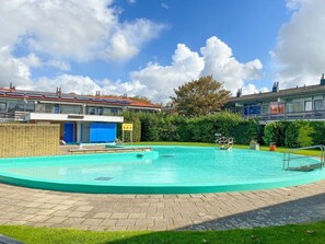 A swimming pool with a brick border, surrounded by a grassy area and a building with a red roof. - Holiday Home With 1 Bedroom (Westkapelle)