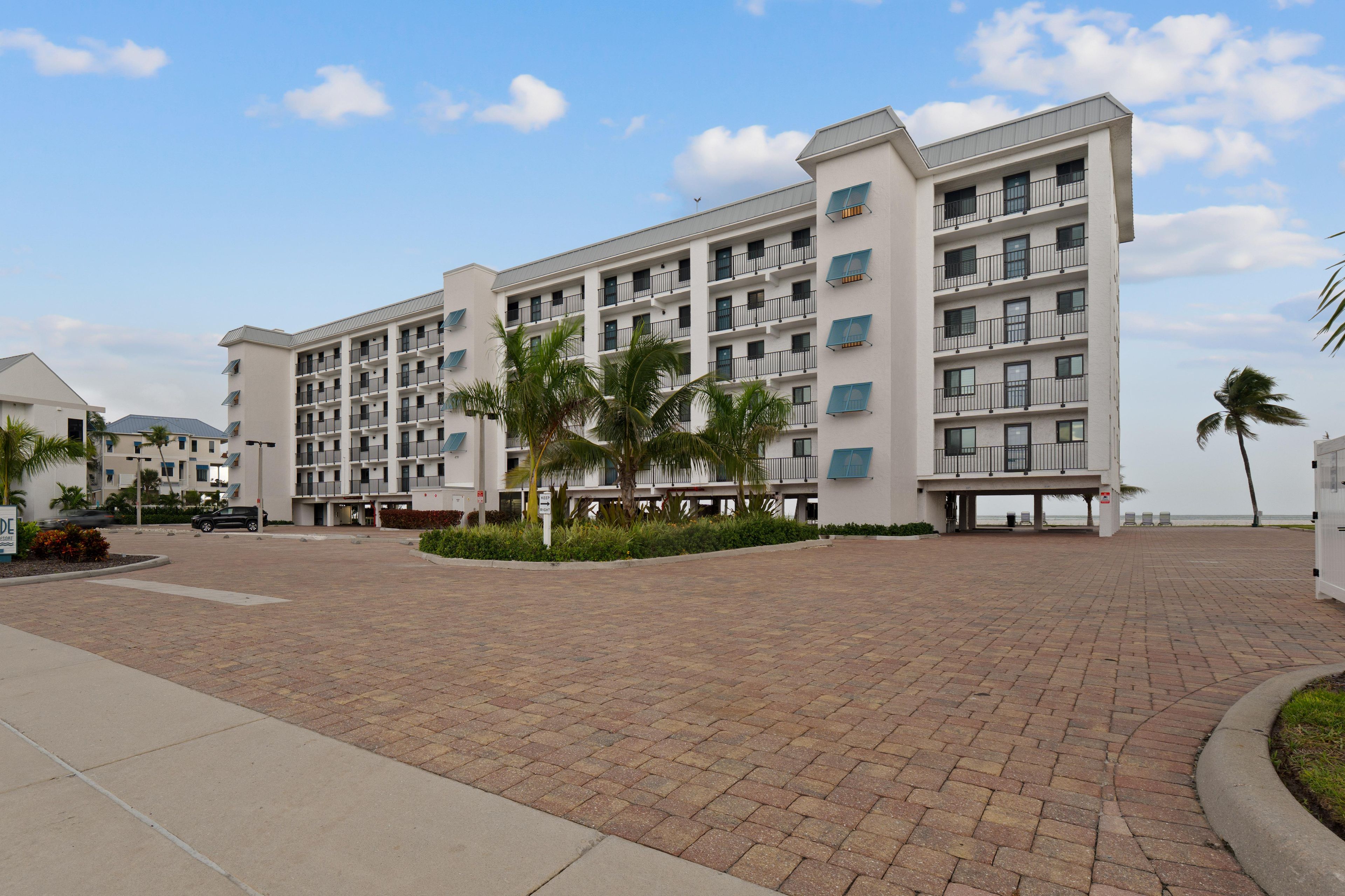 A multi-story hotel with a paved courtyard, palm trees, and a view of the sea.