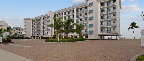 A multi-story hotel with a paved courtyard, palm trees, and a view of the sea.