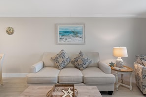 A living room with a beige sofa, two decorative pillows, a framed beach painting, a round wall clock, a side table with a lamp, and a wooden tray with text. - Seaside Condo 109 (Fort Myers Beach)