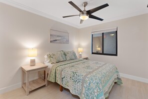A bedroom with a bed, two bedside tables with lamps, a ceiling fan, a painting on the wall, and a window with a view. - Seaside Condo 109 (Fort Myers Beach)