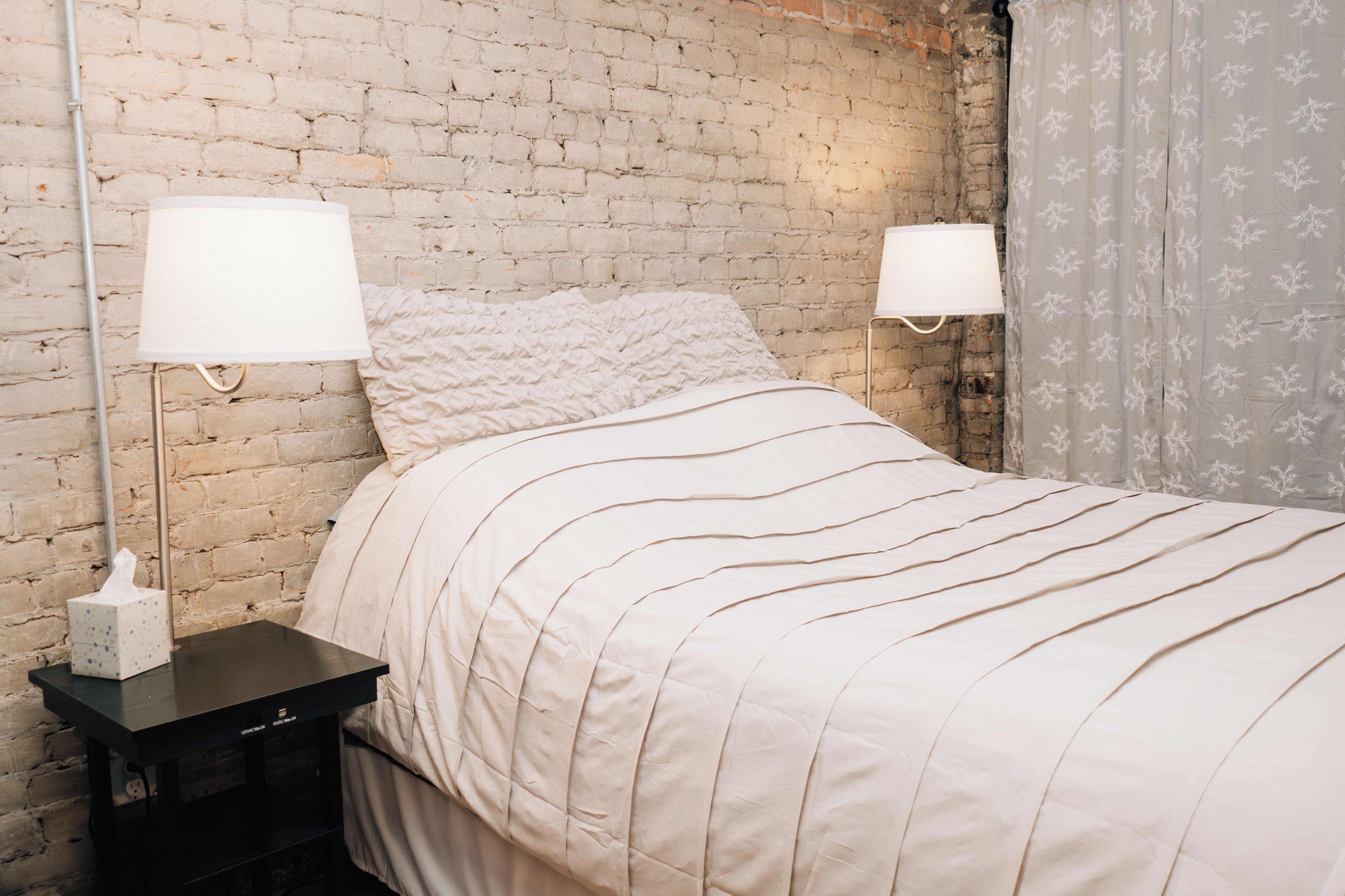 Uma cama com colcha branca, uma mesa de cabeceira com abajur e uma parede de tijolos.