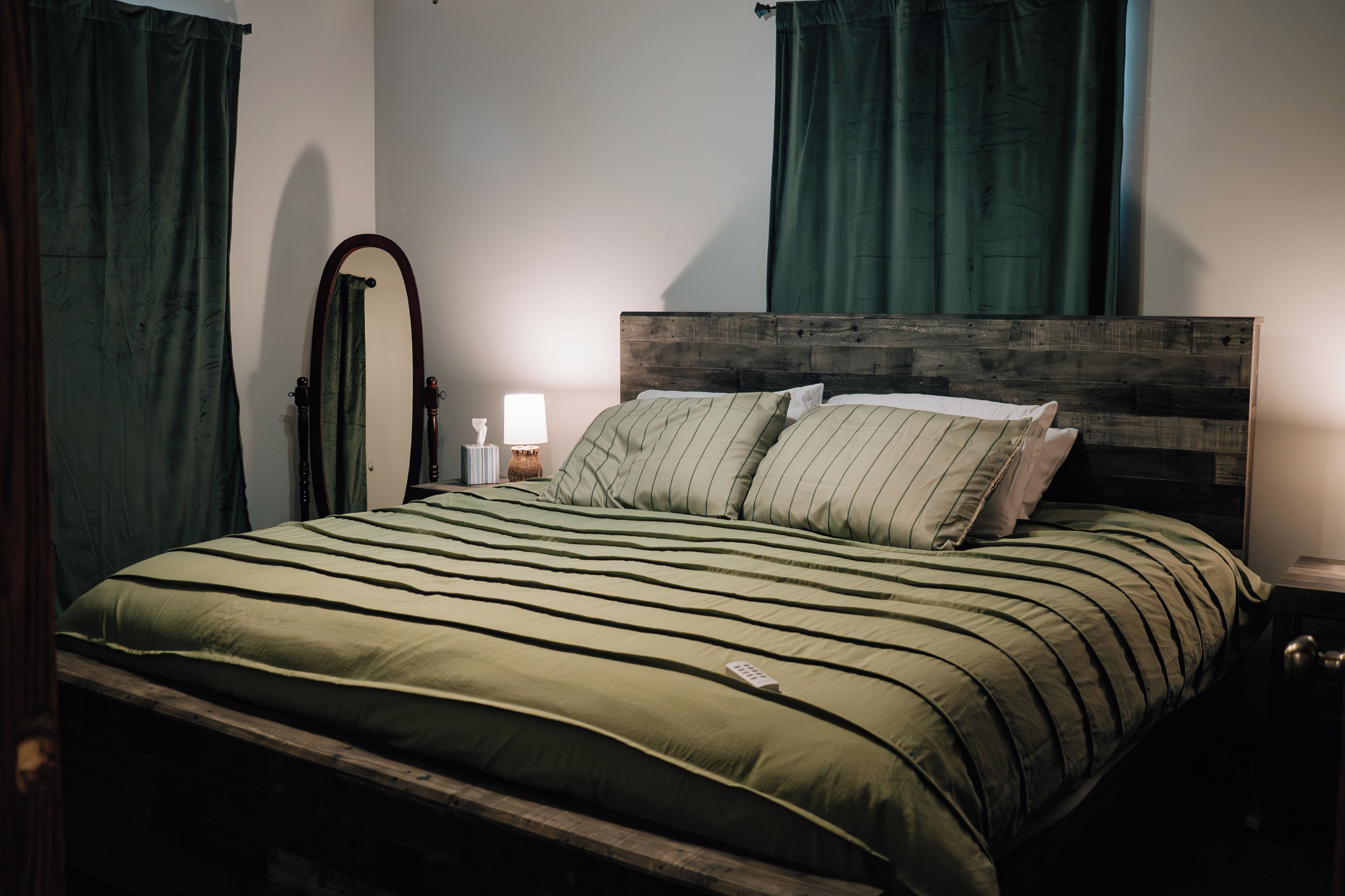 Uma cama com cabeceira de madeira, roupa de cama listrada em verde e mesa de cabeceira com abajur e caixa de lenços.