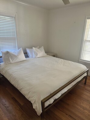 A bedroom with a bed, white bedding, wooden flooring, and a window with blinds. - Georgia’s cottage within walking distance to Texas Tech University. (Lubbock)