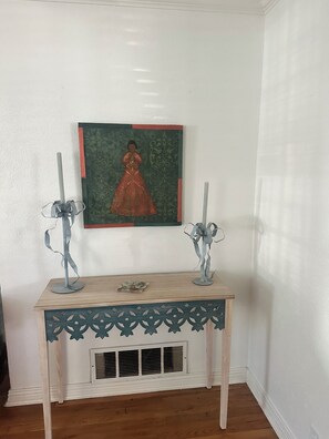 A white wall with a framed painting, a wooden console table with decorative cutouts, and two candle holders with ribbons. - Georgia’s cottage within walking distance to Texas Tech University. (Lubbock)