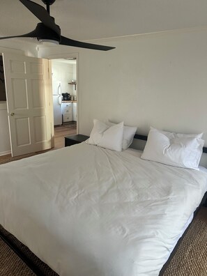 Ein Schlafzimmer mit einem Bett, Kissen, einem Deckenventilator und einer Tür zu einer Küchenzeile.